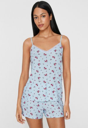 Junge Frau mit langen schwarzen Haaren, die ein hellblaues, geblümtes Camisole und passende Shorts-Pyjamas trägt, steht vor einem einfarbigen Hintergrund.
