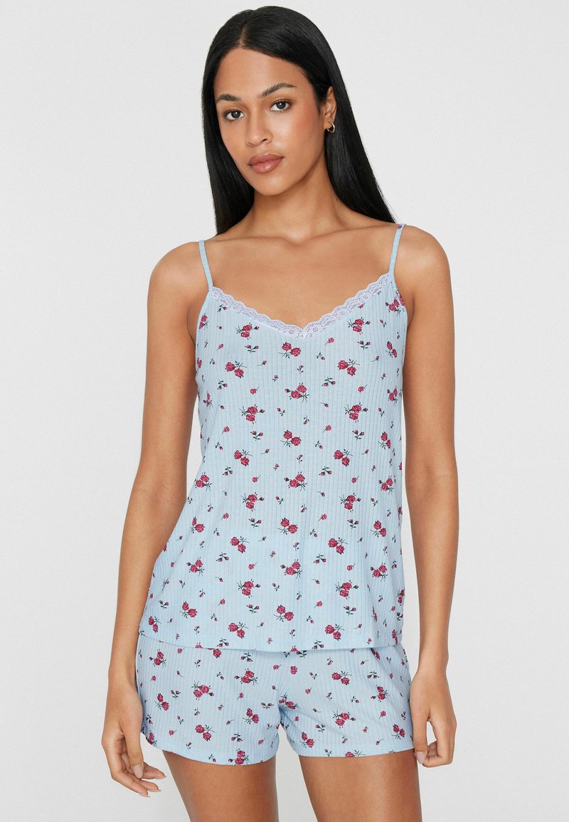 Jeune femme aux longs cheveux noirs portant un débardeur fleuri bleu clair et un pyjama short assorti, debout devant un fond uni.