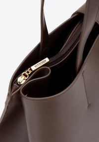 Interno di una borsa a tracolla in pelle marrone con cerniera e accessori dorati. Presenta una texture liscia e un dettaglio inciso "WITTCHEN".
