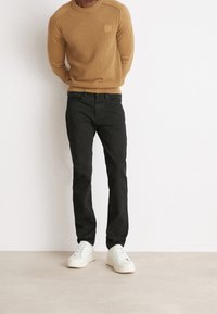 Man vid en vit vägg, iförd en beige tröja, svarta jeans och vita sneakers, står med händerna bakom ryggen.