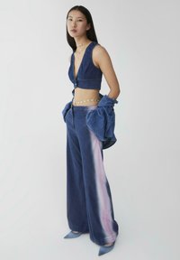 Top corto in denim con scollatura a V profonda, abbinato a pantaloni a gamba larga con una striscia rosa sfumata. Gli accessori includono una cintura in catena dorata e tacchi blu.