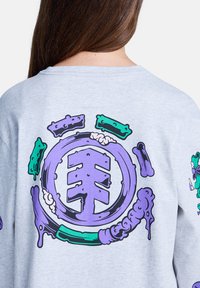 Achteraanzicht van een persoon met lang bruin haar in een lichtgrijze sweatshirt met een kleurrijk paars en teal abstract circulair ontwerp.