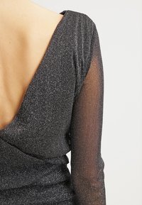 Musta mekko syvällä V-niskalla, jossa on kimaltava kangas, pitkät läpikuultavat hihat ja istuva yläosa, jossa on laskostettu muotoilu vyötäröllä.