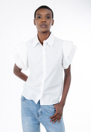 Chemise blanche boutonnée avec manches courtes bouffantes. Tissu lisse, coupe ajustée, col classique et boutons sur le devant. Portée avec un jean bleu.