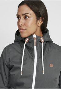 Gray jacket with a hood, white zipper, and contrast drawstrings. Features a brown logo patch and smooth, lightweight material.