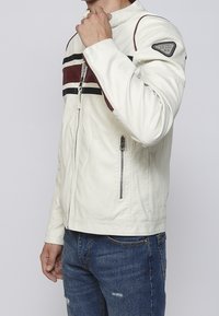 Uomo con giacca bianca con zip, con strisce bordeaux e nere e toppa sulla manica, abbinata a jeans blu.