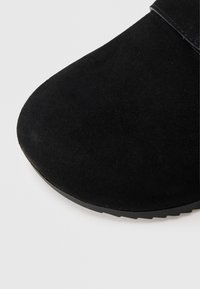 Melns zamšādas apavs ar apaļu purngalu un teksturētu virsmu. Augšējā daļā ir gluda melna siksniņa. Gumijas zole.