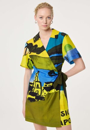 Vrouw draagt een wikkeljurk met korte mouwen en een abstract kleurrijk patroon in geel, blauw, groen en zwart, gestrikt in de taille met een strik.