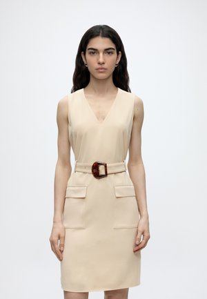 Ung kvinde med langt mørkt hår iført en ærmeløs beige kjole med dyb V-hals, to frontlommer og et bælte med en stor rektangulær spænde.