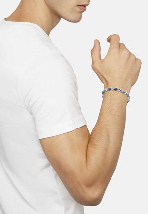 Silbernes Armband mit blauen Akzentperlen, getragen am Handgelenk einer Person in einem weißen T-Shirt, das ein schlankes und abgerundetes Design zeigt.