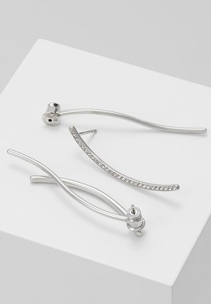 Trois boucles d’oreilles argentées sur une surface blanche : une incurvée avec de petits cristaux, une simple barre incurvée, et un design à double barre torsadée.