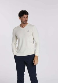 Pull créme en V en tissu doux, avec des manches longues et un petit logo bleu marine sur la poitrine, associé à un pantalon bleu marine.