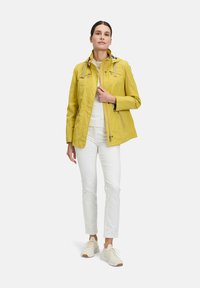 Gelbe wasserdichte Jacke mit Kapuze, Reißverschluss-Taschen und kontrastierenden Reißverschlüssen, getragen über einem weißen Oberteil und weißen Hosen, kombiniert mit hellen Sneakers.
