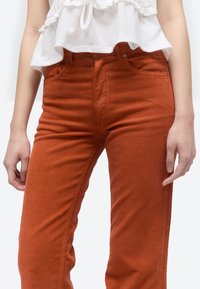 Partea inferioară a torsului poartă pantaloni din catifea de culoare portocaliu ars și un top alb fără mâneci, cu volane, mâinile odihnindu-se pe lângă corp.