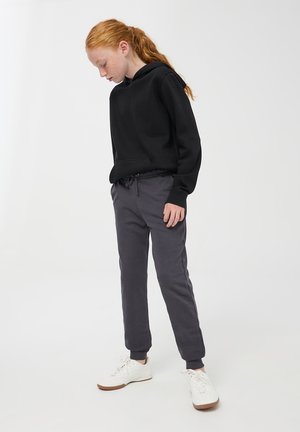 Młoda osoba z rudymi włosami w kucyku, ubrana w czarną bluzę, ciemnoszare dresy i białe trampki, patrząca w dół.