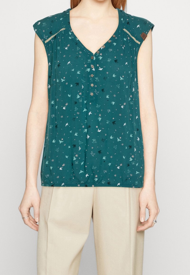 Blouse sans manches couleur sarcelle avec col en V, patte de boutonnage, imprimés d'oiseaux et de fleurs, et pantalon beige porté par une personne du cou aux hanches.