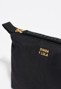 Schwarzes Stofftäschchen mit strukturierter Oberfläche, mit einem goldenen Logo, Reißverschluss und einem schwarzen Tragegurt mit eingraviertem "BIMBA Y LOLA".