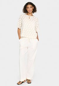 Blusa in pizzo color crema con maniche corte e colletto con fiocco, abbinata a pantaloni bianchi larghi e ciabatte nere. Tessuto morbido e vestibilità rilassata.