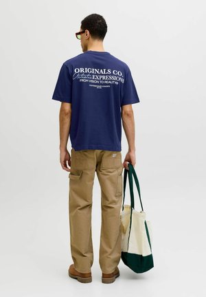 Mann trägt marineblaues Grafik-T-Shirt und beige Cargohose, hält weiße und grüne Tragetasche, steht mit dem Rücken zu einfarbigem Hintergrund.