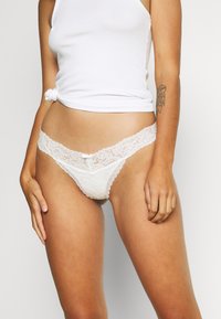 Witte kanten string met een scalloped kant tailleband, een kleine centrale strikdetail en gladde stof voor de kern. Zachte en delicate textuur.