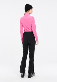 Maglione in pile rosa a maniche lunghe abbinato a pantaloni da neve neri con dettagli in zip alle caviglie, indossato con stivali da sci neri.