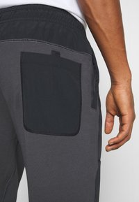 Close-up van de hand van een persoon naast donkergrijze broek met een zwart patch zak en een elastische tailleband.