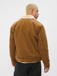 Brun cordfløyeljakke med en kremfarget faux pelskrage. Har sidelommer, knappede mansjetter og et teksturert stoff. Rent design.