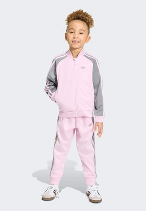 Jeune enfant debout, les mains dans les poches, portant un survêtement Adidas rose et gris ainsi que des baskets blanches sur un fond uni.