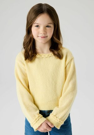 Niña joven con cabello castaño ondulado, vistiendo un suéter amarillo con detalles trenzados y jeans azules, de pie con las manos entrelazadas.