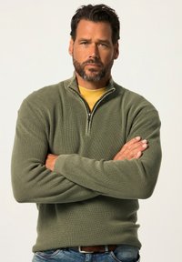 Olijfgroene fleece-jas met geribbelde textuur, lange mouwen en een opstaande kraag, gedragen over een gele T-shirt.