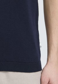 Cămașă tricotată bleu marin cu margine ribbed și detalii; prim-planul arată textura moale și un mic logo alb pe lateral.