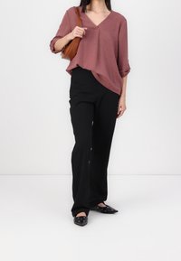 Roestkleurige, lichte V-hals blouse met opgerolde mouwen, gecombineerd met zwarte wijde broek en zwarte instap schoenen met zilveren accenten.