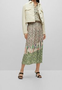 Krémová džínsová bunda s kapsami na hrudi, spárovaná s maxi šaty s mnoha vzory, které obsahují květinové a geometrické designy v pastelových barvách.