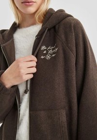 Bruine fleece zip-up hoodie met een kangoeroezak, geborduurde tekst "Wees vriendelijk & Gelukkig" en een verstelbare trekkoordcapuchon. Zachte textuur.