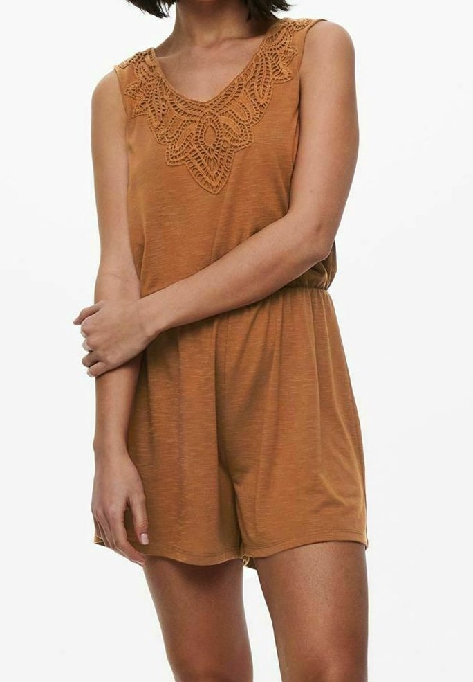 Combinaison sans manches de couleur orange rouille avec un détail en dentelle sur l'encolure en V, une taille élastique et un short ample portée par une personne debout, les bras croisés.