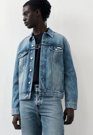 Mladý muž s tmavou pokožkou a dredmi, oblečený v svetlomodrej rifľovej bunde a zodpovedajúcich rifliach, pozerajúci sa doľava na jednoliatom pozadí.