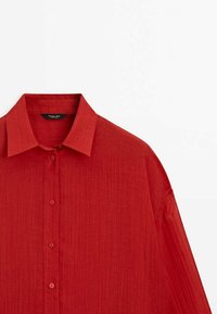 Chemise rouge à boutons avec un col et des manches longues, présentant un tissu texturé et de discrètes rayures verticales.