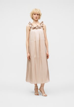 Γυναίκα με σγουρά ξανθά μαλλιά που φοράει αμάνικο, παστέλ ροζ φόρεμα μέτριου μήκους με βολάν στον λαιμό και μπεζ ψηλοτάκουνα σανδάλια, όρθια.