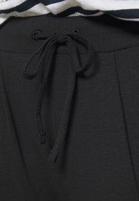 Pantalon de jogging noir avec une texture douce, doté d'un cordon à la taille et d'un design plissé. En arrière-plan, une chemise rayée.