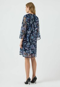 Vestido de encaje floral azul marino con mangas transparentes, con bordados intrincados en azul y plata. Dobladillo con borde festoneado y ajuste a medida. Zapatos de tacón altos negros.