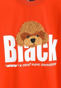 Camiseta de algodón roja con un aplique de un perro marrón texturizado en el frente. Un texto blanco en negrita dice "Black" encima de un logo de marca más pequeño.