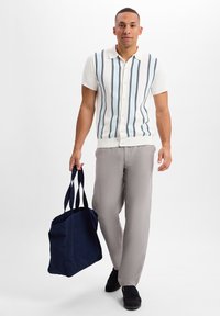 Kurze weiße Strickbluse mit navyblauen vertikalen Streifen, kombiniert mit hellgrauen Hose. Hält eine dunkelblaue Canvas-Tasche.