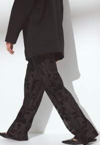 Melni caurspīdīgi bikses ar ziedu rakstu, teksturētu virsējo slāni, plata kāju dizains, saskaņotas ar brīvu melnu blūzi un melniem apaviem.