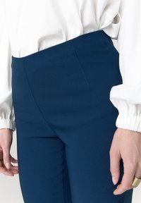 Marineblauwe, nauwsluitende broek met een gladde textuur, hoge taille en zijnaaddetails, gecombineerd met een witte top met ballonmouwen.