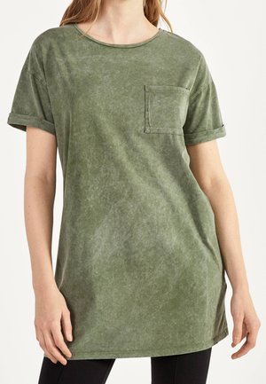 Kobieta ubrana w luźny, krótki zielony bawełniany t-shirt z kieszenią na piersi i podwiniętymi rękawami, zestawiony z czarnymi spodniami.
