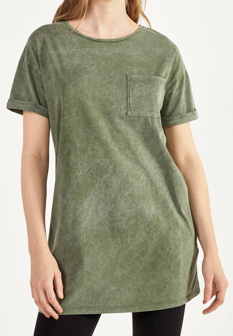 Femme portant un T-shirt ample à manches courtes en coton vert lavé, avec une poche poitrine et des manches retroussées, assorti à un pantalon noir.
