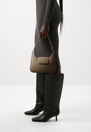 Mujer con vestido oscuro acanalado y botas negras de tacón hasta la rodilla, sosteniendo un bolso de cuero marrón por su asa superior.