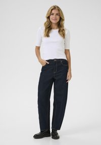 Jeans de denim azul oscuro con una cintura alta y piernas anchas, con costuras naranjas y diseño clásico de cinco bolsillos, combinados con una blusa blanca ajustada.