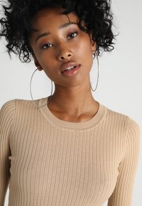 Top en tricot côtelé beige avec un col rond. Doté de manches longues et d'une texture douce. De grandes boucles d'oreilles en argent complètent le look.