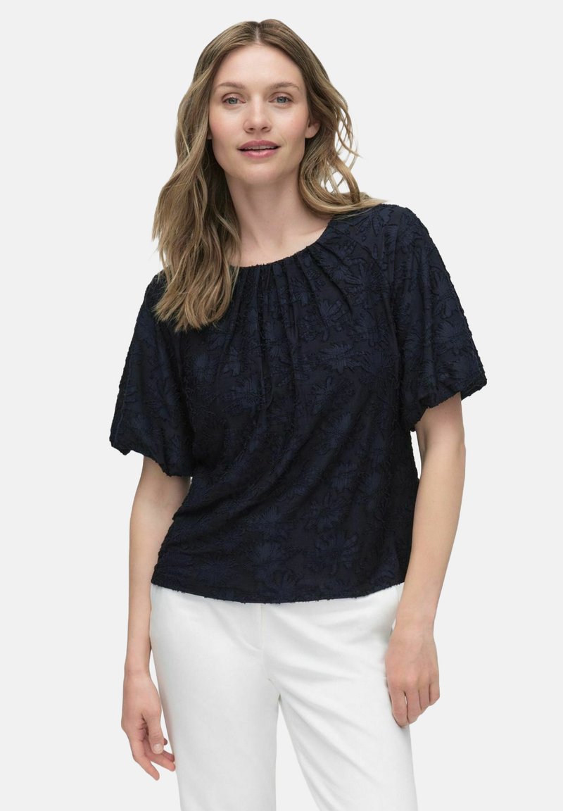 Navy blå kortærmet bluse med tekstureret blomsterdesign, rund halsudskæring, blødt stof og afslappet pasform, parret med hvide bukser.